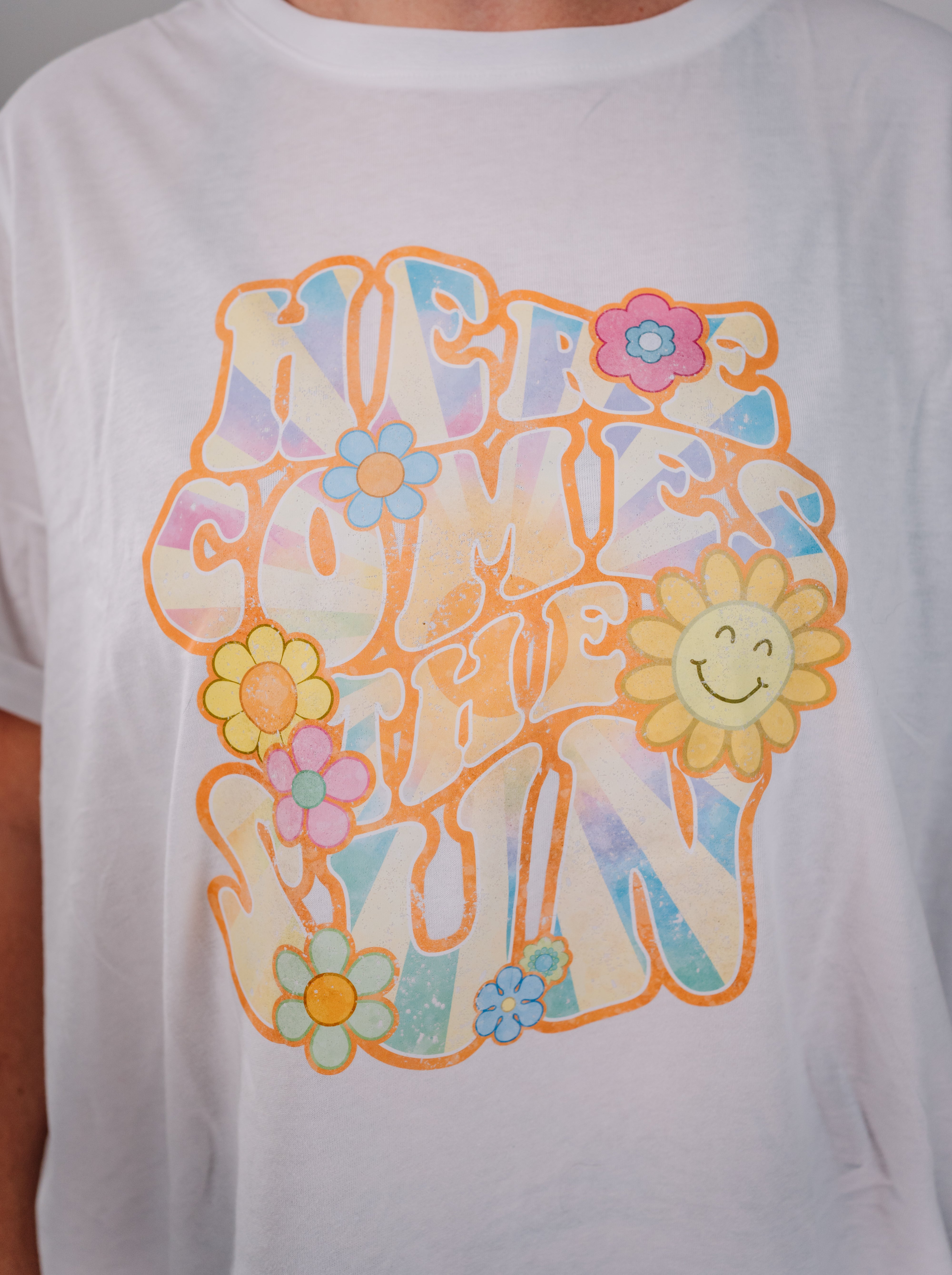 A white short-sleeve women's t-shirt with a colorful graphic print that includes the text 'HERE COMES THE SUN' surrounded by sunflowers and a smiling sun face.