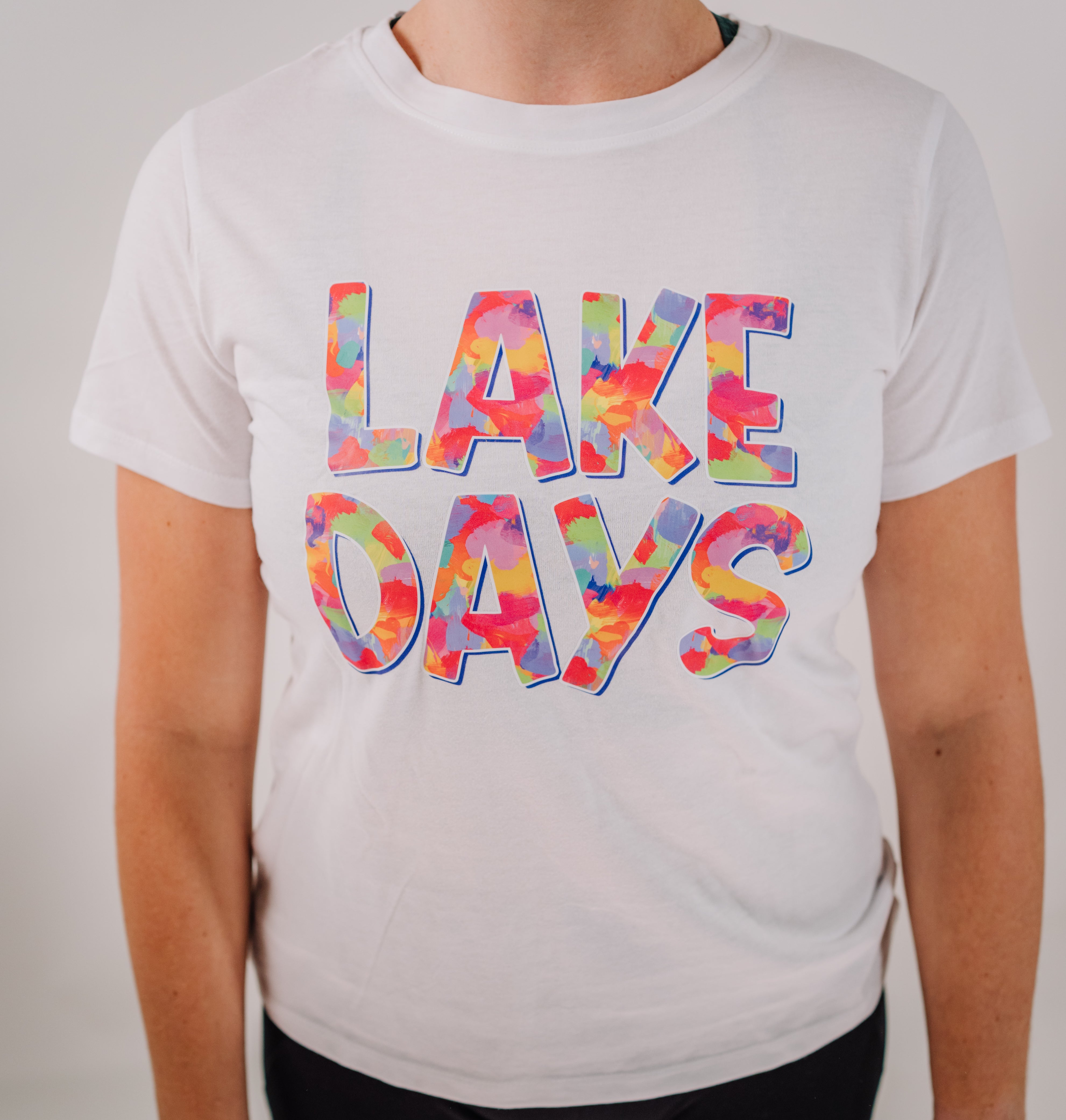 A woman wearing a white short sleeve t-shirt with the text 'LAKE DAYS' printed across the front in a colorful, gradient-style design.