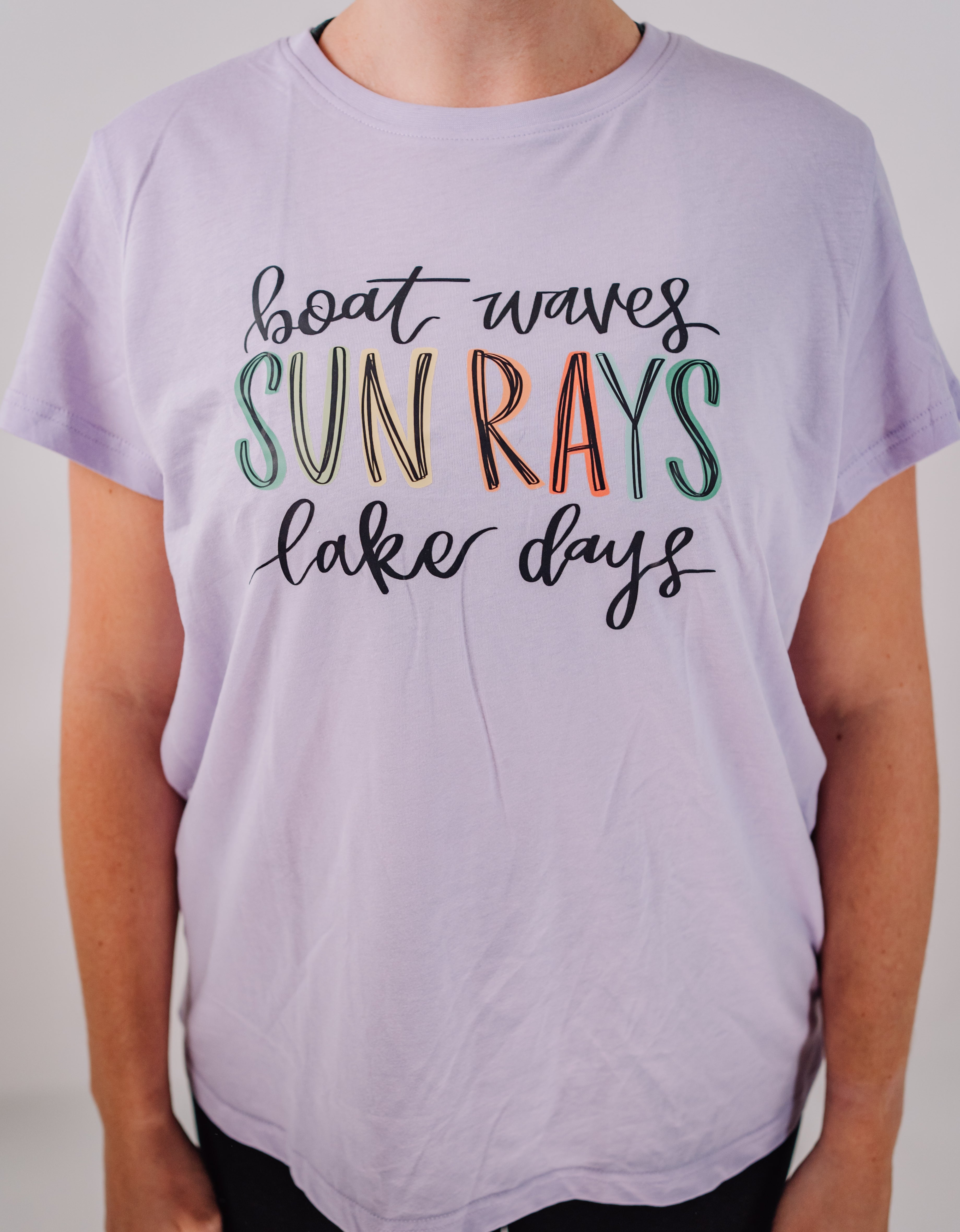 A person wearing a short sleeve women's t-shirt in purple color with the text 'boat waves SUN RAYS lake days' printed on it in a multicolor font.