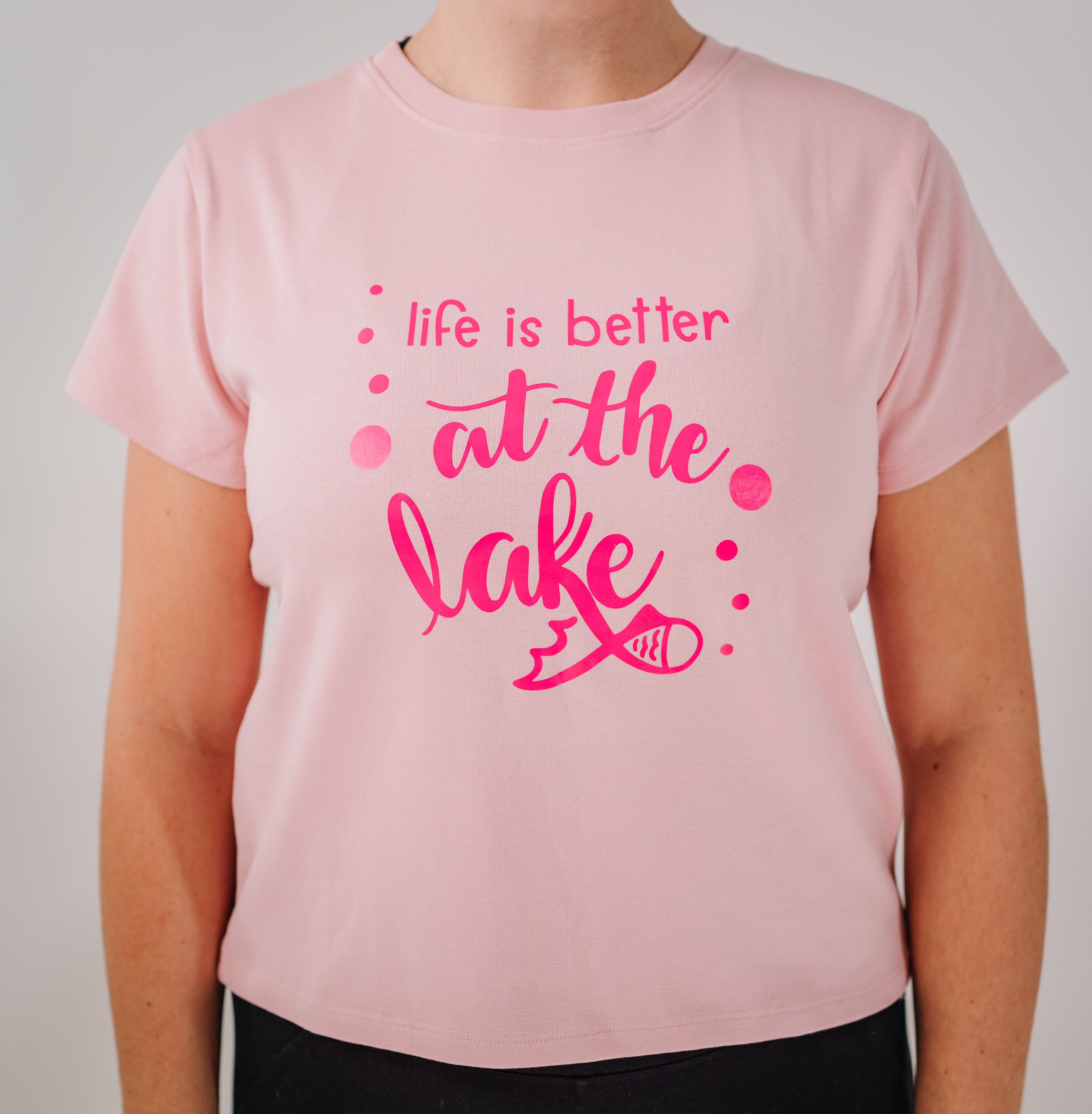 A pink short-sleeved women's t-shirt with the text 'LIFE IS BETTER AT THE LAKE' printed on the front.